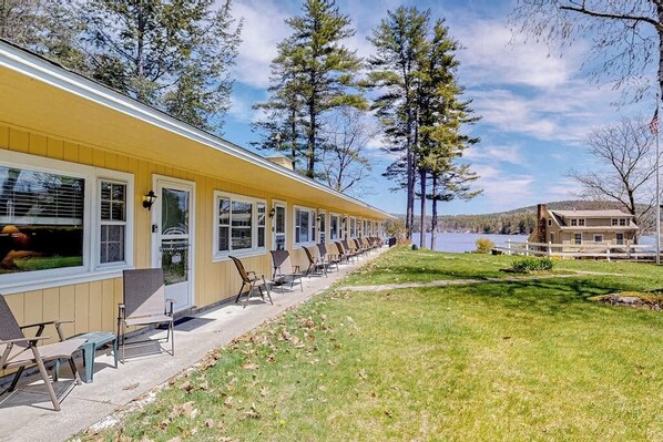 A row of single-story yellow buildings with large windows, outdoor seating, and a view of a lake.
