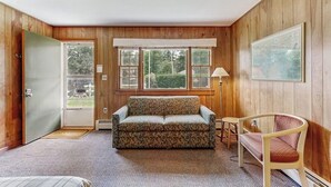 Ein Zimmer mit Holzwänden, einer Couch, einem Sessel, einem kleinen Tisch und einem Fenster mit Blick auf Grünflächen.