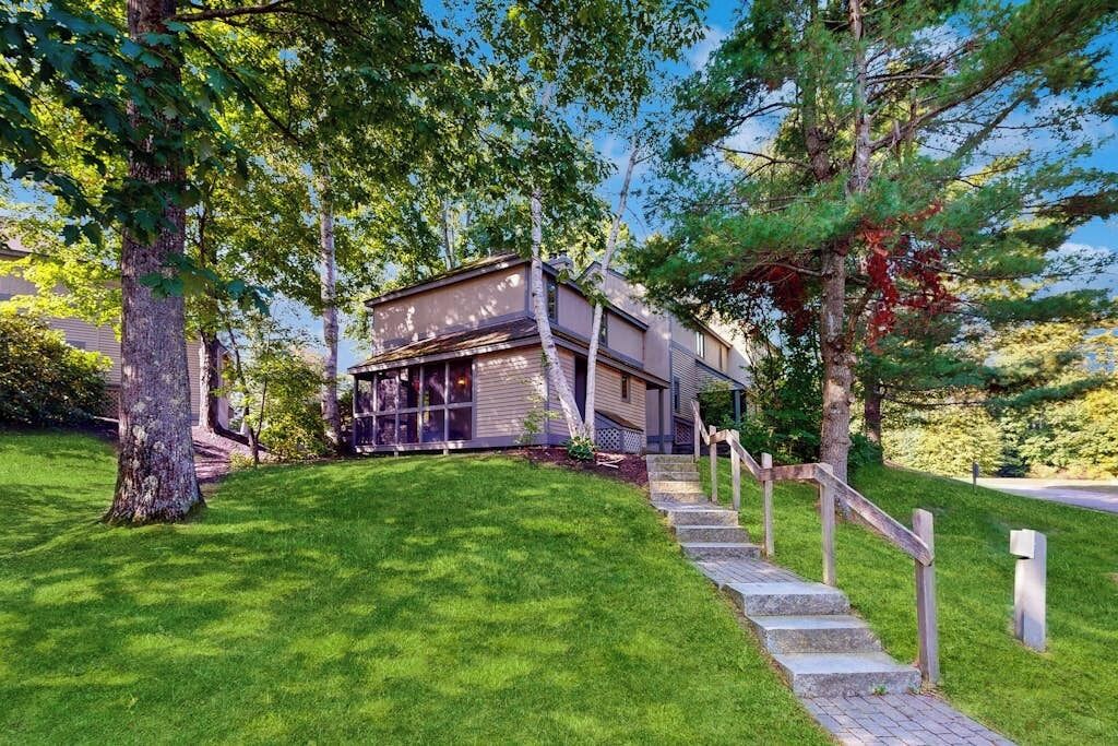 A modern two-story house with a stone staircase leading to the front door, surrounded by lush green grass and trees.