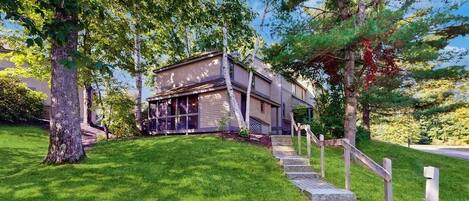 A modern two-story house with a stone staircase leading to the front door, surrounded by lush green grass and trees.