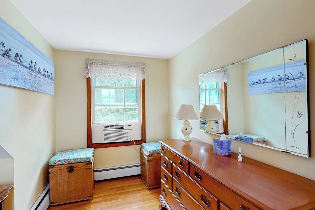 A room with a wooden dresser, a bench, a window with a lace curtain, and wall-mounted artwork.