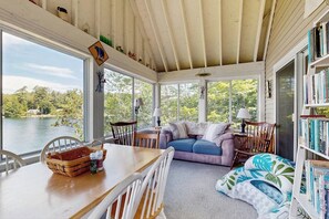 A sunroom with a view of a lake, a dining table with chairs, a sofa, and a bookshelf. - Scenic 2BR Lakefront - Washer Dryer (Meredith)