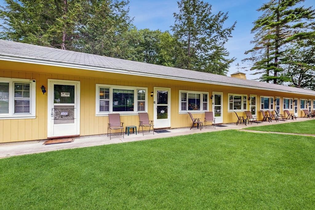 Une rangée de chambres d’un motel jaune, toutes de plain-pied, avec des fenêtres individuelles, un espace extérieur abrité avec des sièges et une pelouse bien entretenue.