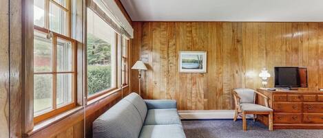 A room with a wooden paneled wall, a sofa, a desk with a television, and a window with blinds.