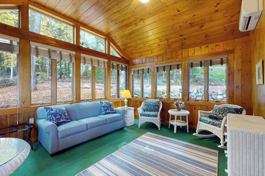 Ein gemütliches Blockhaus-Interieur mit Holzbalkendecke, großen Fenstern, einem blauen Sofa, Korbstühlen und einem Streifen Teppich.