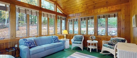 Ein gemütliches Blockhaus-Interieur mit Holzbalkendecke, großen Fenstern, einem blauen Sofa, Korbstühlen und einem Streifen Teppich.