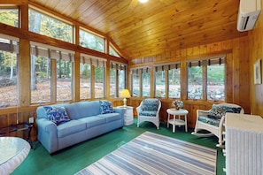 A cozy cabin interior with a wooden ceiling, large windows, a blue sofa, wicker chairs, and a striped rug. - Stunning 1BR Lakefront - Dock - Deck (Moultonborough)
