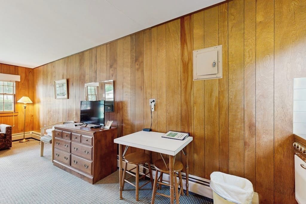 Une pièce aux murs lambrés, avec un bureau blanc et deux chaises, une télévision, une commode et une fenêtre.