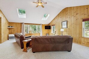 Ein gemütliches Zimmer mit einem braunen Sofa, einem Holztisch, einem Deckenventilator und einem an der Wand befestigten Fernseher.