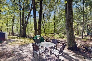 Eine Holzterrasse mit rundem Tisch und Stühlen, umgeben von Bäumen in einem bewaldeten Gebiet.