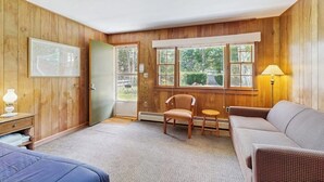 Quarto com painéis de madeira, uma cama, uma poltrona, um sofá e uma janela com vista para o verde.