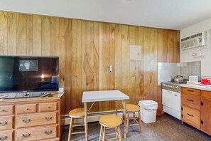 A compact kitchen with wooden paneling, a television, a small dining table, two stools, a white stove, and a trash bin. - Convenient Studio Lakefront - Kitchenette (Alton)