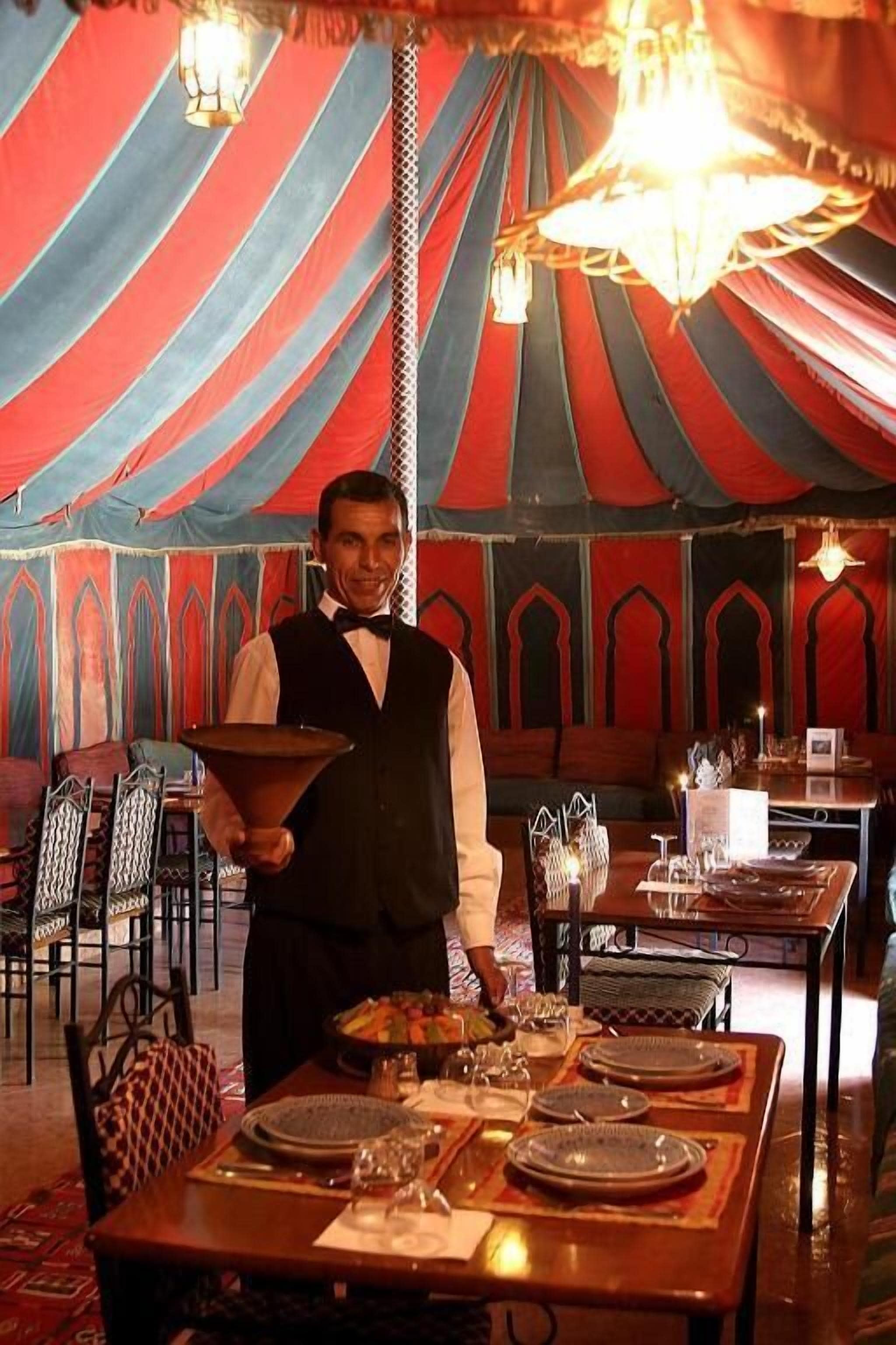A dining area with a tent-like structure, tables set with plates and glasses, and a waiter holding a bowl.