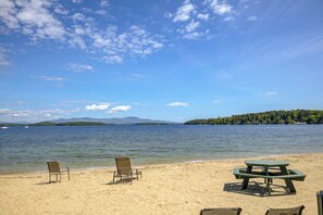 A beach with a picnic table and two chairs, a clear sky, and a lake with a forested island in the distance. - Conveniently located 1BR 2nd-Floor - Deck (Gilford)