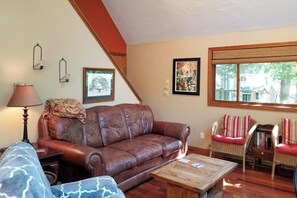 A living room with a brown leather sofa, a wooden coffee table, a lamp, and a window with a view of trees. - 3BR Near the Beach - Deck - Tennis - Washer Dryer (Moultonborough)