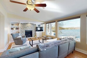 Ein Wohnzimmer mit Kamin aus Stein, einem Fernseher, einer Couch und Blick auf ein Gewässer.