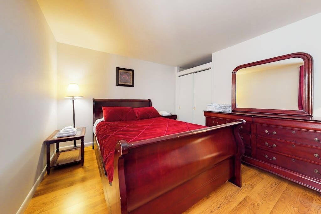 Een slaapkamer met een houten bed, een nachtkastje, een lamp, een dressoir en een spiegel.