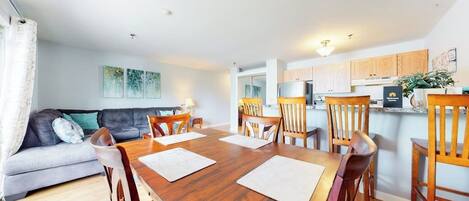 A modern living room with a dining table and chairs, a sofa, and a kitchen area with wooden cabinets.