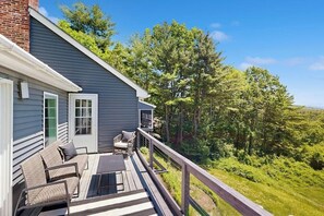 A deck with outdoor furniture, a brick chimney, and a view of a forested landscape. - 3BR stunning home with views, pool & lake access (Meredith)