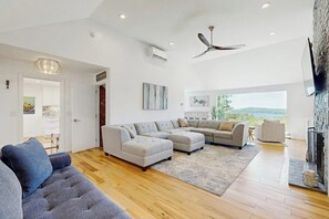 A spacious living room with a sectional sofa, a flat-screen TV mounted on the wall, and a large window offering a view of the outdoors. - 3BR stunning home with views, pool & lake access (Meredith)