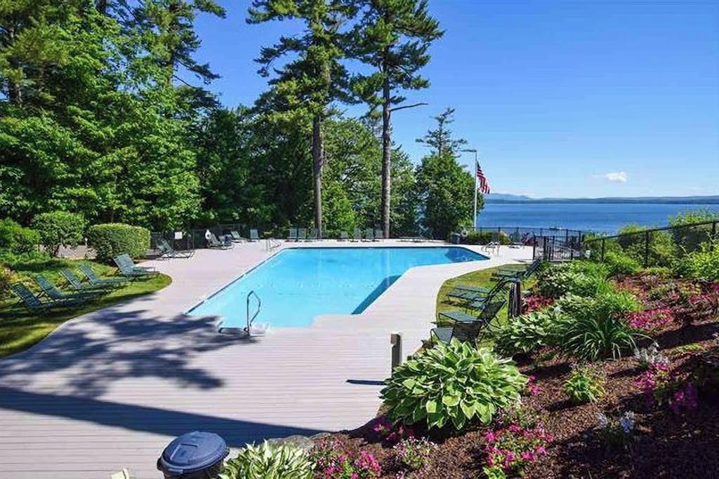 A swimming pool with a wooden deck, surrounded by greenery and a view of the water.
