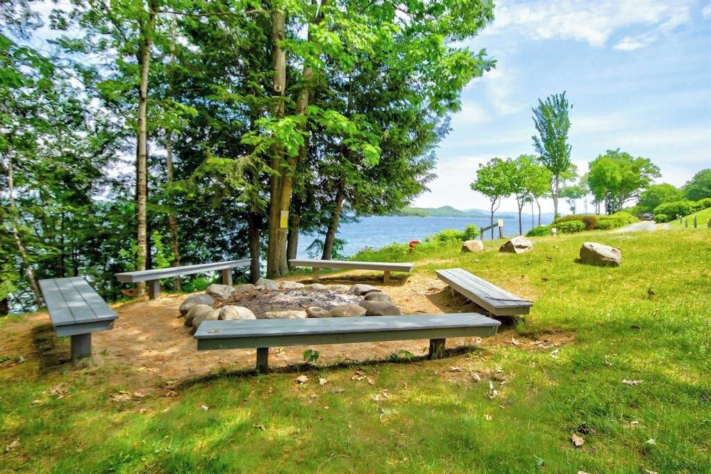 A lakeside picnic area with benches and a fire pit surrounded by trees.