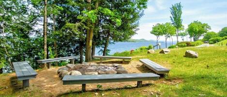 A lakeside picnic area with benches and a fire pit surrounded by trees.