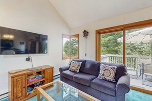 A living room with a sofa, coffee table, wooden cabinet with board games, a mounted TV, and large windows with a view of greenery. - 3BR withbeach, pool, tennis, game room, porch &views (Gilford)