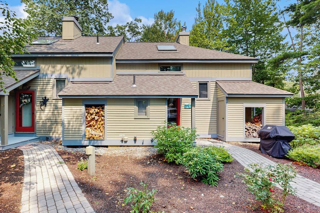 Ein zweistöckiges Haus mit überdachter Veranda, einem Holzstapel und einem Grill im Garten.