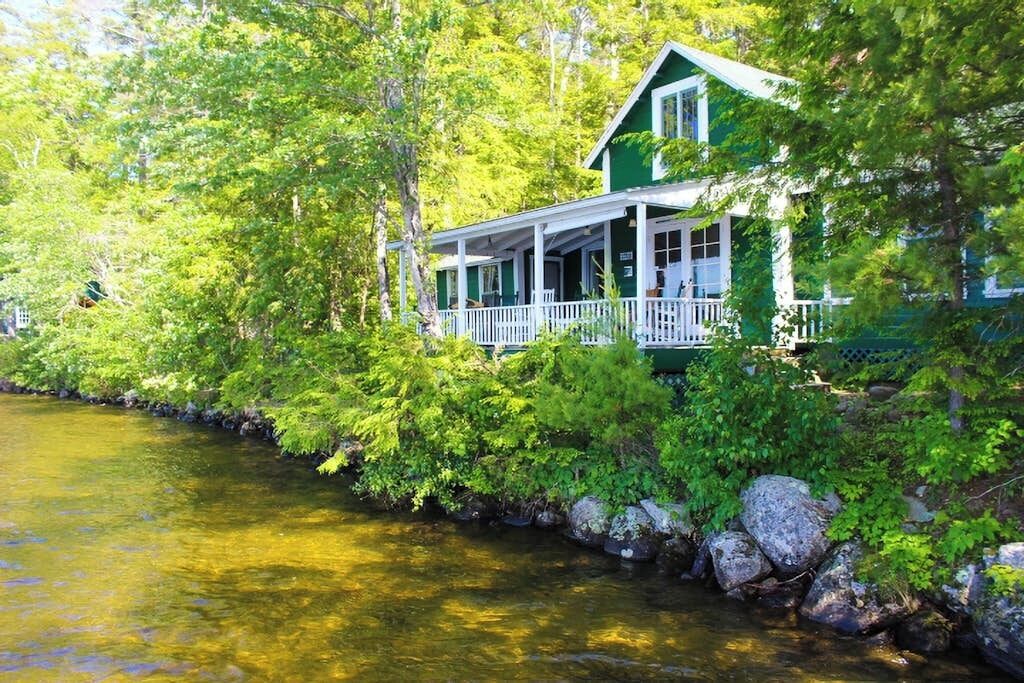 A green house with a porch overlooking a river, surrounded by dense greenery.