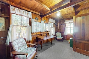 Interior de uma cabana de madeira com teto de madeira, mesa de jantar, duas cadeiras, um sofá e uma janela com cortinas.