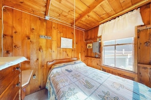 Ein Zimmer einer Holzhütte mit einem Bett, einem Schminktisch, einem Fenster mit Vorhängen und Blick ins Freie.