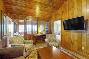 A wooden-paneled room with a television mounted on the wall, a wooden table, and patterned armchairs. - Stunning 2BR Lakefront - Dock - Patio - Washer Dryer (Moultonborough)