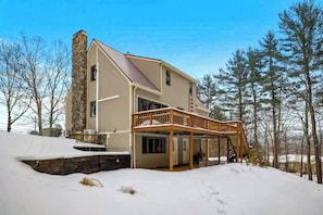 A two-story house with a stone chimney, a covered porch, and a snow-covered landscape. - 3BR home near outdoor rec with deck & games (Gilford)