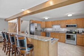 A kitchen with wooden cabinets, a granite countertop, stainless steel appliances, and a bar area with stools. - 3BR home near outdoor rec with deck & games (Gilford)