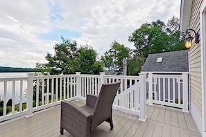 A wooden deck with a white railing, a wicker chair, and a view of a lake and trees. - Lake view 2BR with dock & deck, walk to the beach (Moultonborough)