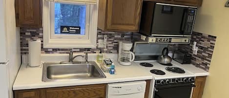 A compact kitchen with wooden cabinets, a built-in microwave, a stove, a dishwasher, and a window with a view of trees.