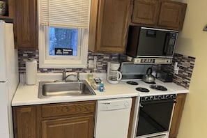 A compact kitchen with wooden cabinets, a built-in microwave, a stove, a dishwasher, and a window with a view of trees. - 1BR Lakeview 2nd-Floor - Balcony - Outdoor Pool (Gilford)