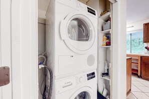 A laundry area with stacked washing machines, a visible power outlet, and a view of a kitchen area with cabinets and a window. - Serene 3BR Dog Friendly - Sauna - Balcony (Gilford)