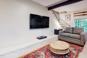 Ein modernes Wohnzimmer mit einem an der Wand befestigten Flachbildfernseher, einem grauen Sofa, einem Holztisch und einem gemusterten Teppich.