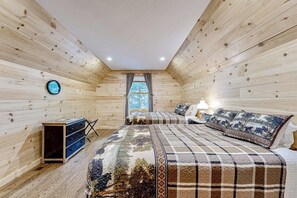 Una habitación con paredes de madera, una cama con colcha estampada, un aparador y una ventana con cortinas.