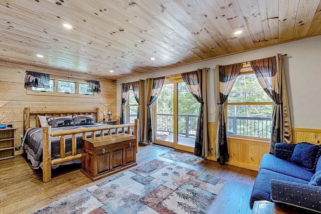A wooden cabin interior with a bed, a sofa, and a large window with curtains.