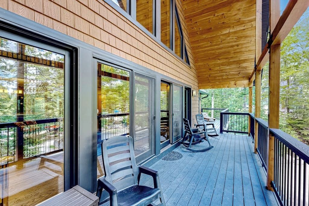 A wooden deck with a covered seating area, large windows, and a view of the surrounding forest.
