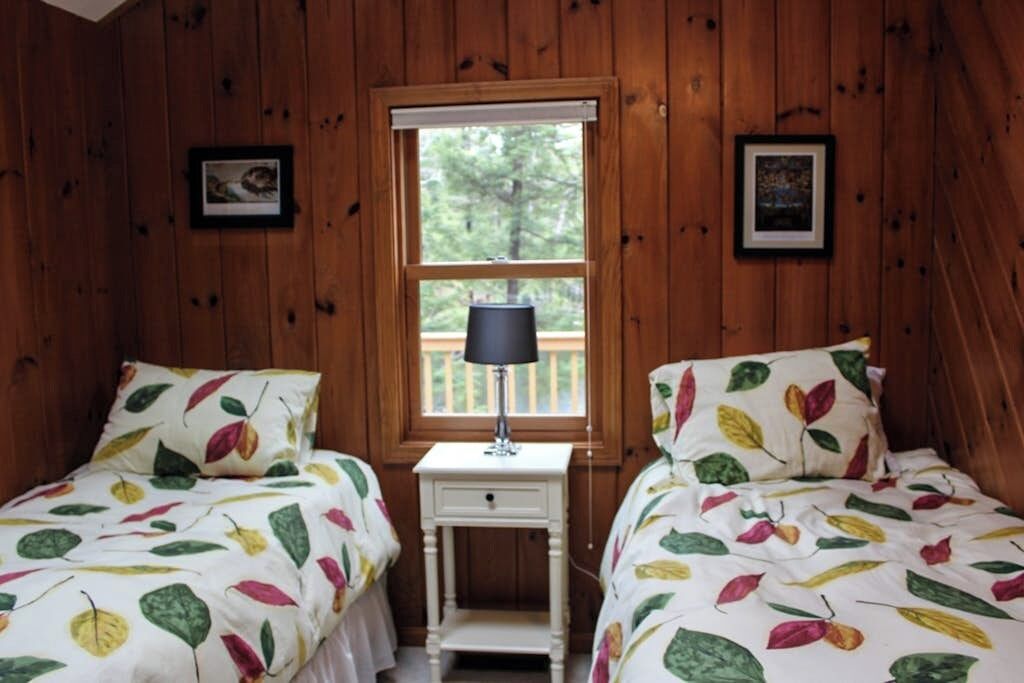 Une pièce aux murs en bois, avec deux lits dotés de draps à motifs de feuilles, une petite table de chevet blanche avec une lampe, et une fenêtre donnant sur des arbres.