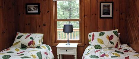 Una habitación con paredes de madera, dos camas con ropa de cama estampada con motivos de hojas, una mesita de noche blanca pequeña con una lámpara y una ventana con vistas a árboles.