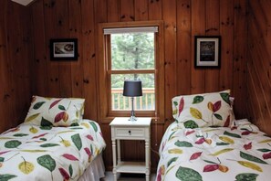Een kamer met houten wanden, twee bedden met beddengoed met een bladmotief, een klein wit nachtkastje met een lamp, en een raam met uitzicht op bomen.