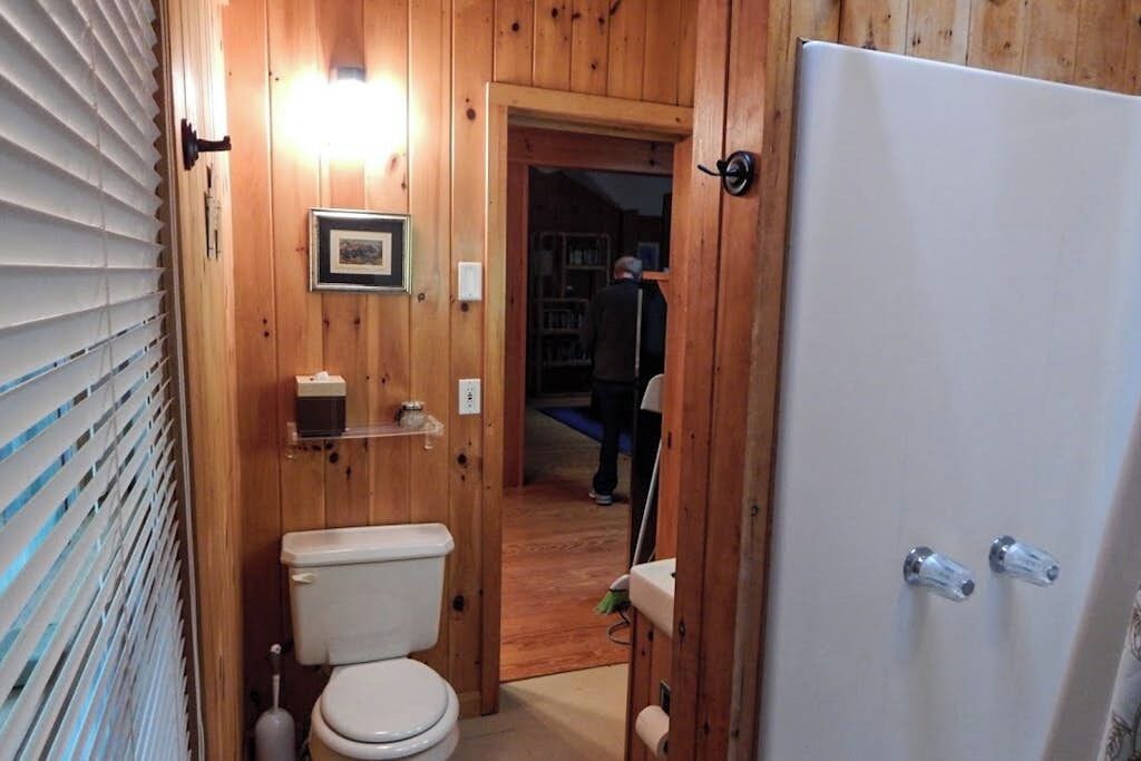 Un baño con paneles de madera, un inodoro, un estante con un objeto pequeño y una puerta que conduce a otra habitación.