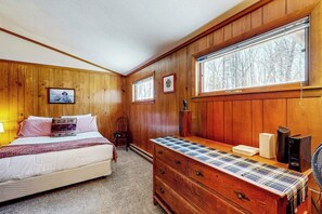 Ein Schlafzimmer mit einer holzverkleideten Wand, einem Bett mit einer karierten Tagesdecke, einer Kommode mit einem gemusterten Deckel und einem Fenster mit Blick auf Bäume.