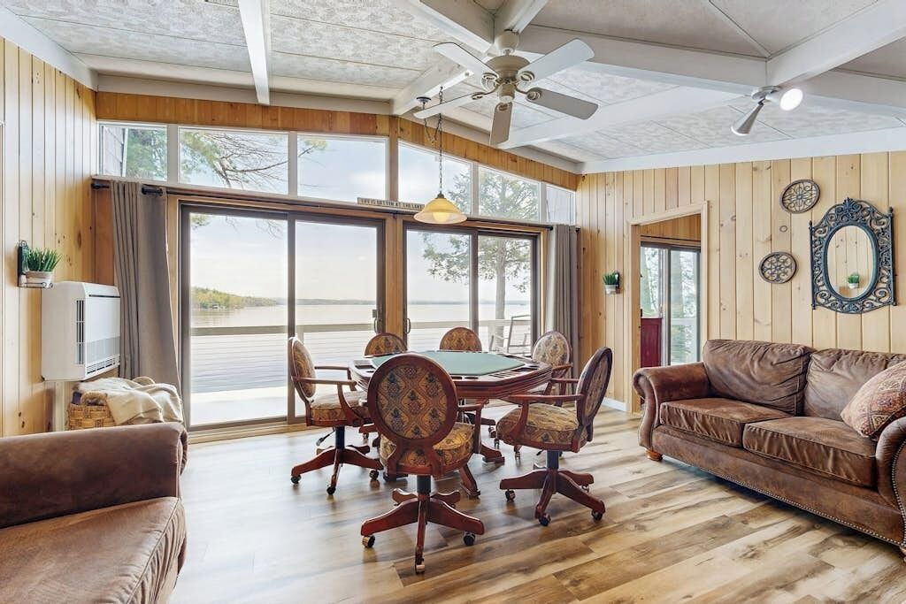 Una habitación amplia con un gran comedor, pisos de madera, un ventilador de techo y ventanas grandes.