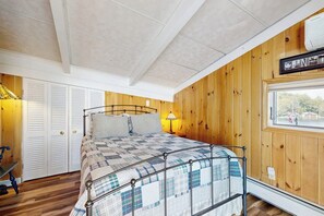 Une chambre à coucher avec un cadre de lit en métal, une couvre-lit matelassée, une fenêtre avec vue, des murs en bois et un ventilateur de plafond.